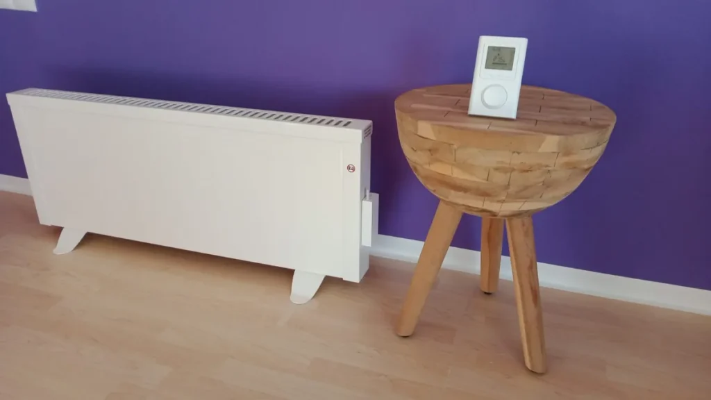 Weiße Elektroheizung neben einem Hocker mit digitalem Thermostat vor violetter Wand